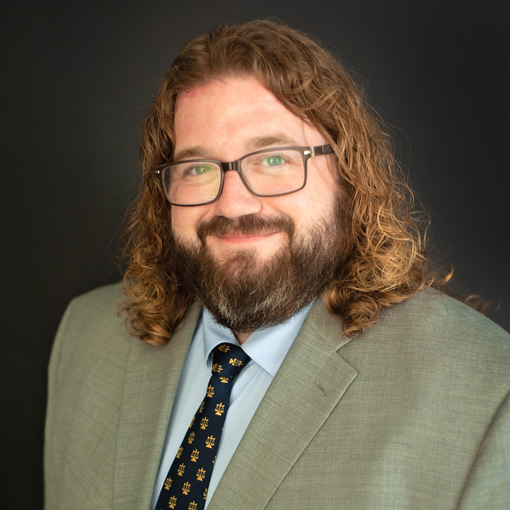 A person with long curly hair and a beard wears glasses, a light gray suit, a blue shirt, and a patterned tie. The background is solid black.