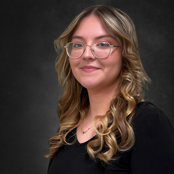 A woman with long, wavy hair and glasses is smiling against a dark background. She is wearing a black top and a delicate necklace.