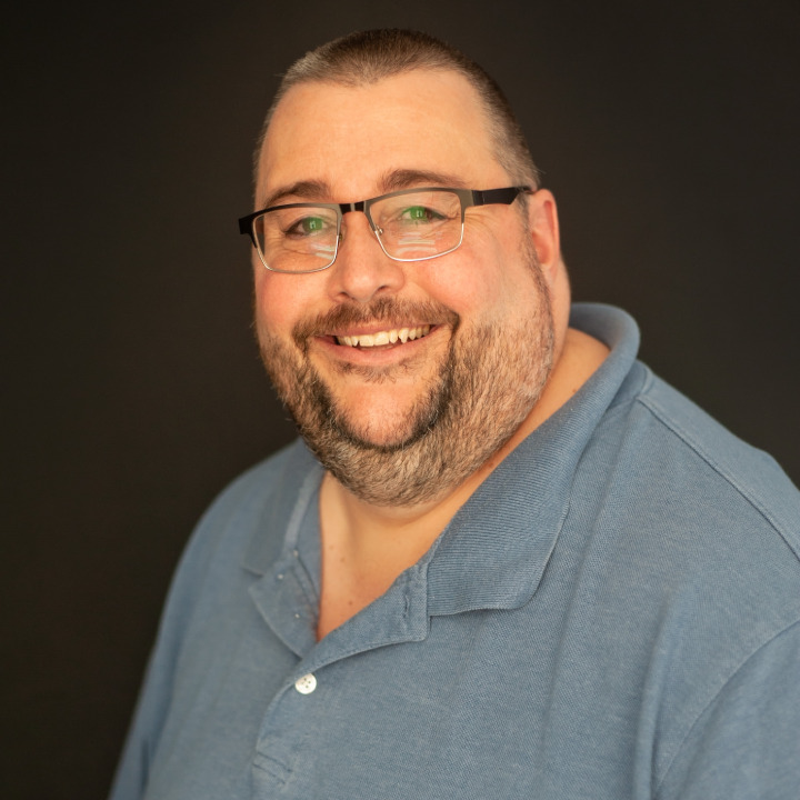 Smiling man wearing glasses and a blue polo shirt in front of a dark background.