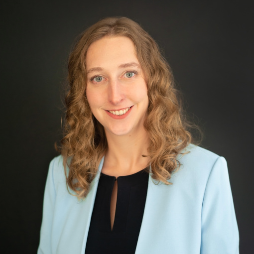 A woman with curly light brown hair smiles at the camera. She is wearing a light blue blazer over a black top, set against a plain dark background.