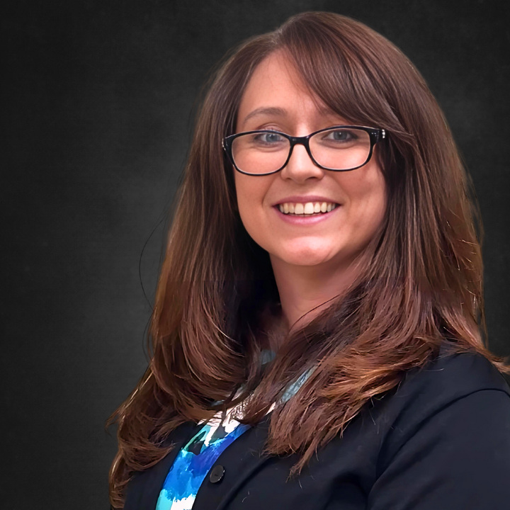 A woman with long brown hair wearing glasses and a dark blazer smiles against a dark background.