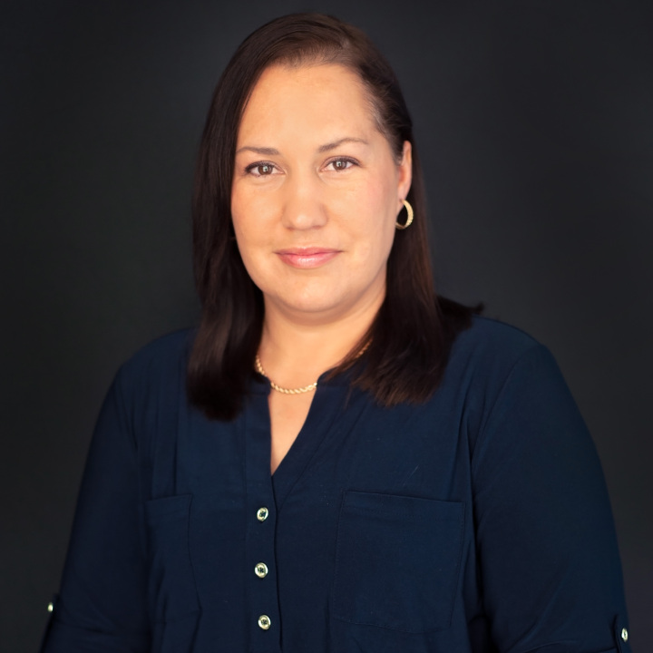 A person with shoulder-length dark hair is wearing a dark blue shirt and gold earrings, smiling subtly against a plain dark background.