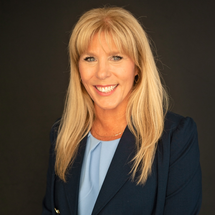 Smiling woman with long blonde hair wearing a dark blue blazer and light blue top against a plain dark background.