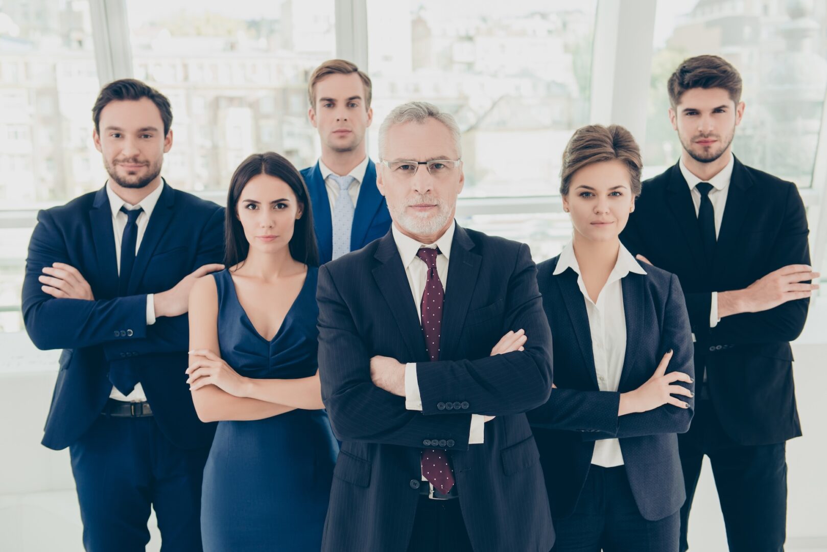 A group of six business professionals stands confidently in an office setting. The older man in the center wears glasses and a suit, flanked by three men and two women, all dressed in formal business attire with arms crossed.