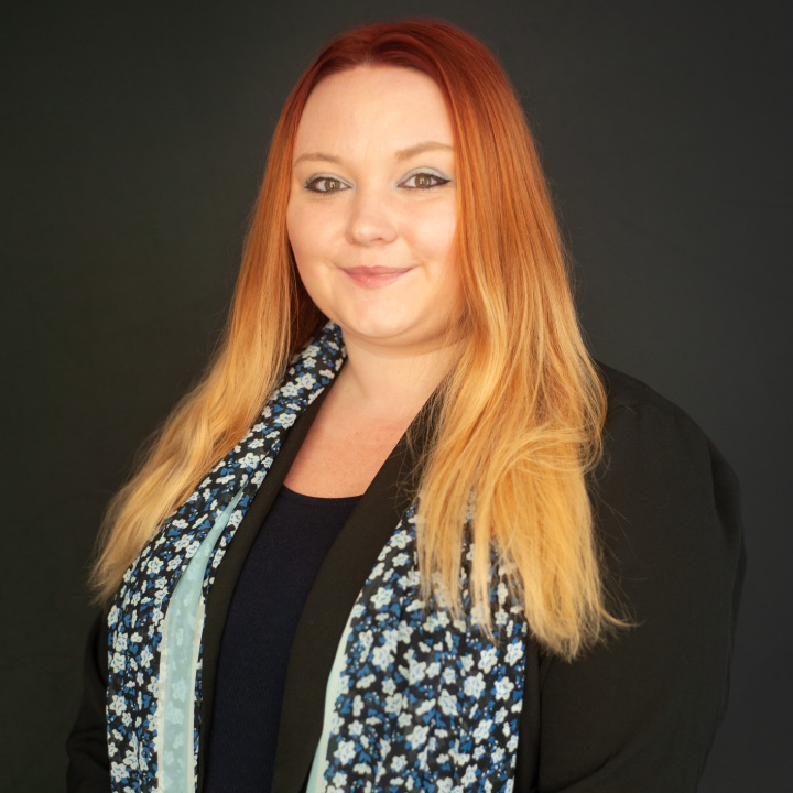 A person with long, flowing red hair smiles warmly while wearing a black jacket and a light blue scarf with floral patterns, standing against a dark background.