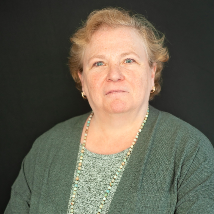 A woman with short blonde hair, wearing a gray cardigan and patterned top, looks directly at the camera. The background is solid dark.