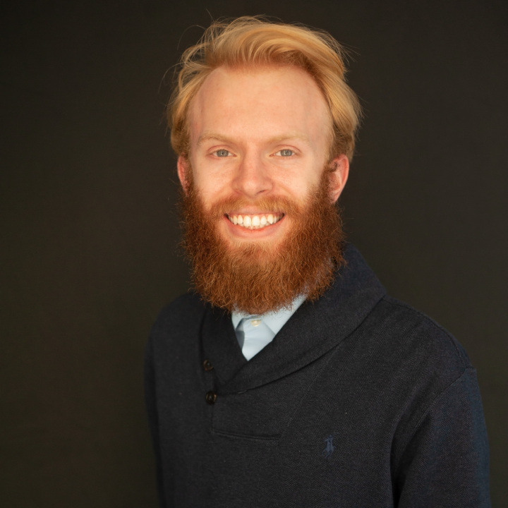 A person with red hair and a beard smiles at the camera. They're wearing a dark sweater over a light shirt. The background is a plain, dark color.