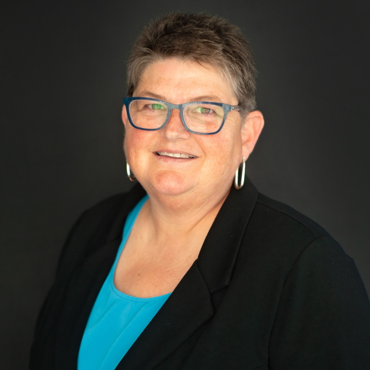 A person with short hair, wearing glasses and hoop earrings, smiles at the camera. They are dressed in a black blazer over a blue top, set against a plain dark background.