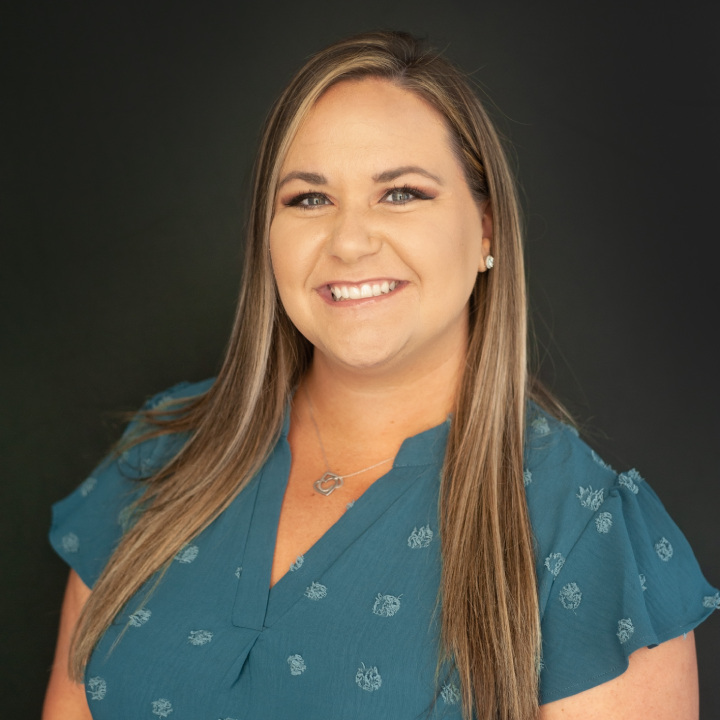 A woman with long, straight hair smiles at the camera. She is wearing a teal blouse with a pattern and a heart-shaped necklace. The background is dark, highlighting her face.