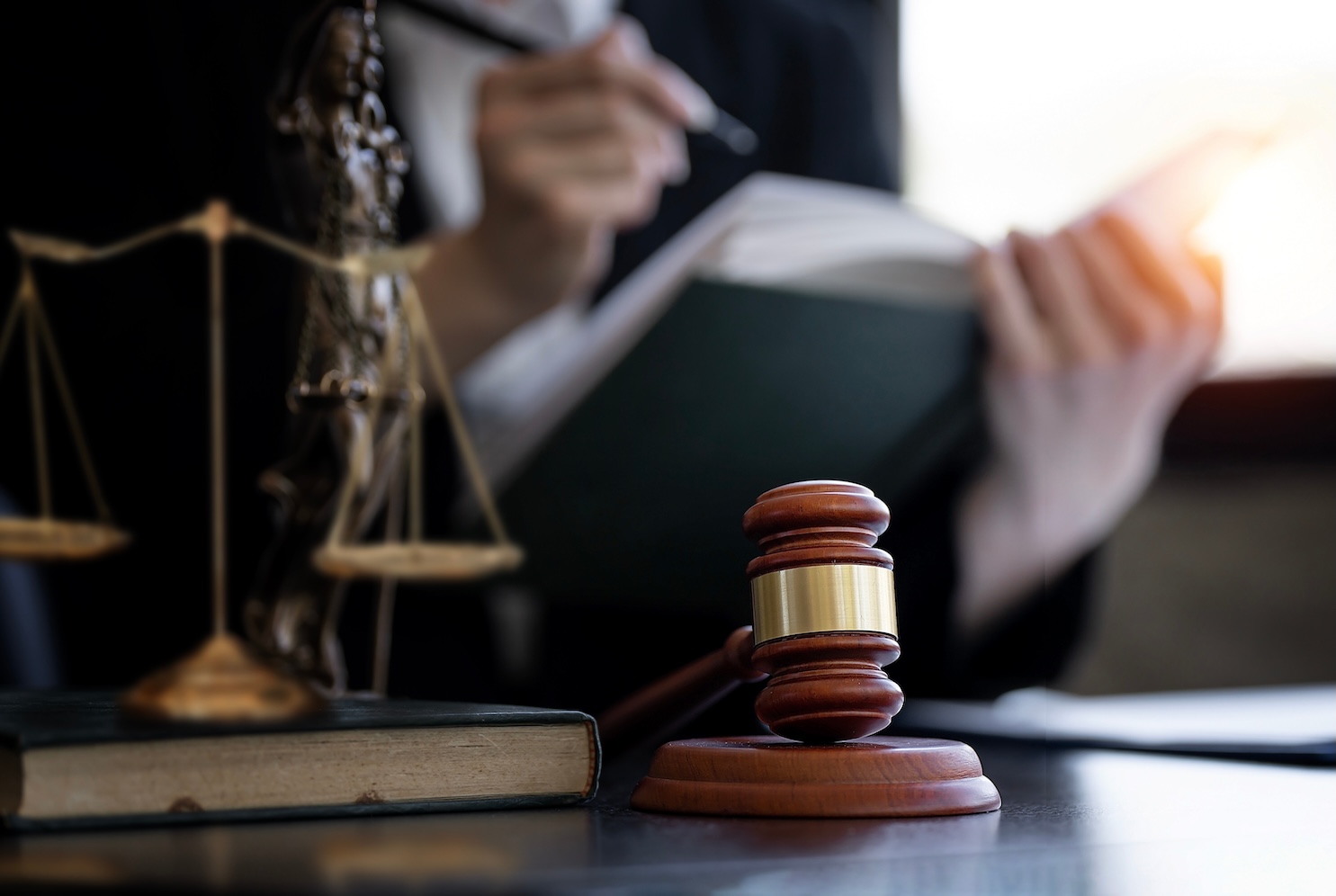 A wooden gavel and scales of justice on a desk, with a person in a suit writing in a book in the background. The scene is lit by warm lighting, creating a professional and serious atmosphere.