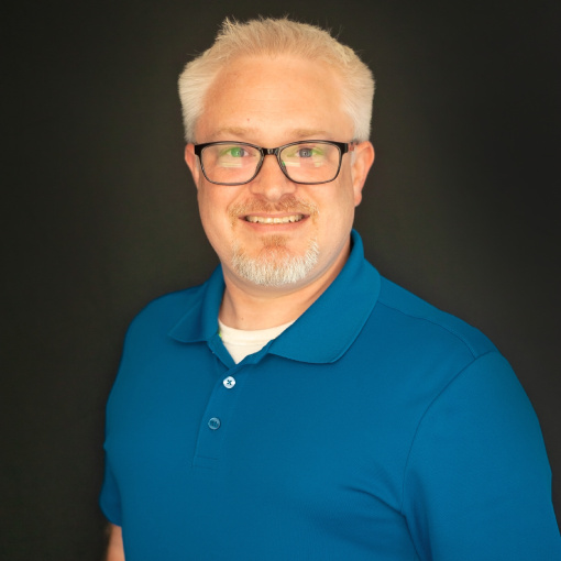 A person with short, light-colored hair and glasses is smiling at the camera. They are wearing a blue polo shirt against a dark background.