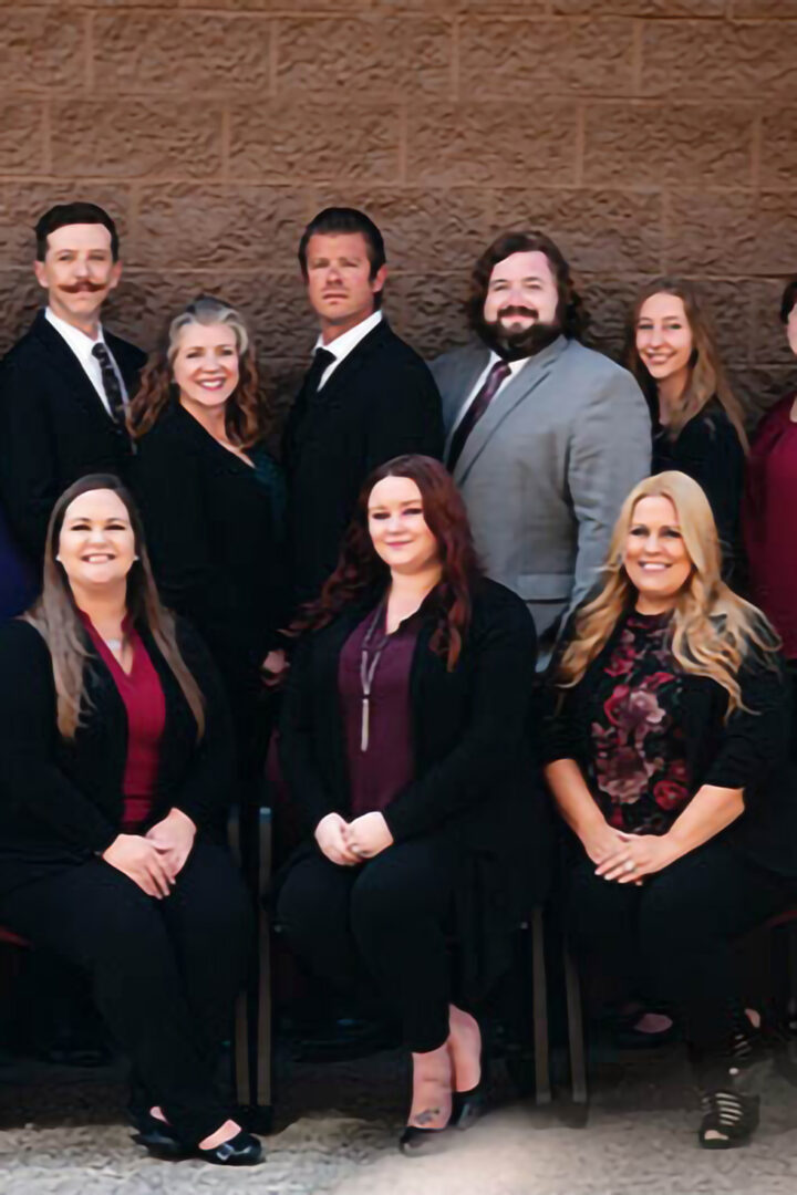 A group of nine people, dressed in business attire, are posing for a formal photo against a brown textured wall. Some are standing while others are seated in the front row, all smiling.