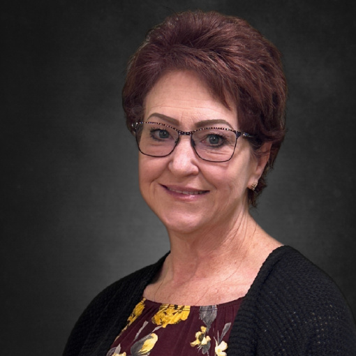 A woman with short brown hair and glasses smiles warmly. She is wearing a floral blouse and a dark cardigan. The background is a plain, dark gray.