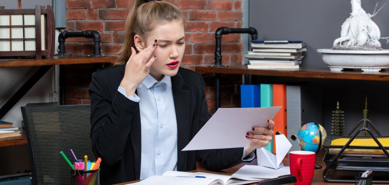 A woman in a suit sits at a desk, looking frustrated while holding a piece of paper. The desk is cluttered with stationery, books, a globe, and a red cup. She is touching her temple with her other hand. The background has a brick wall.