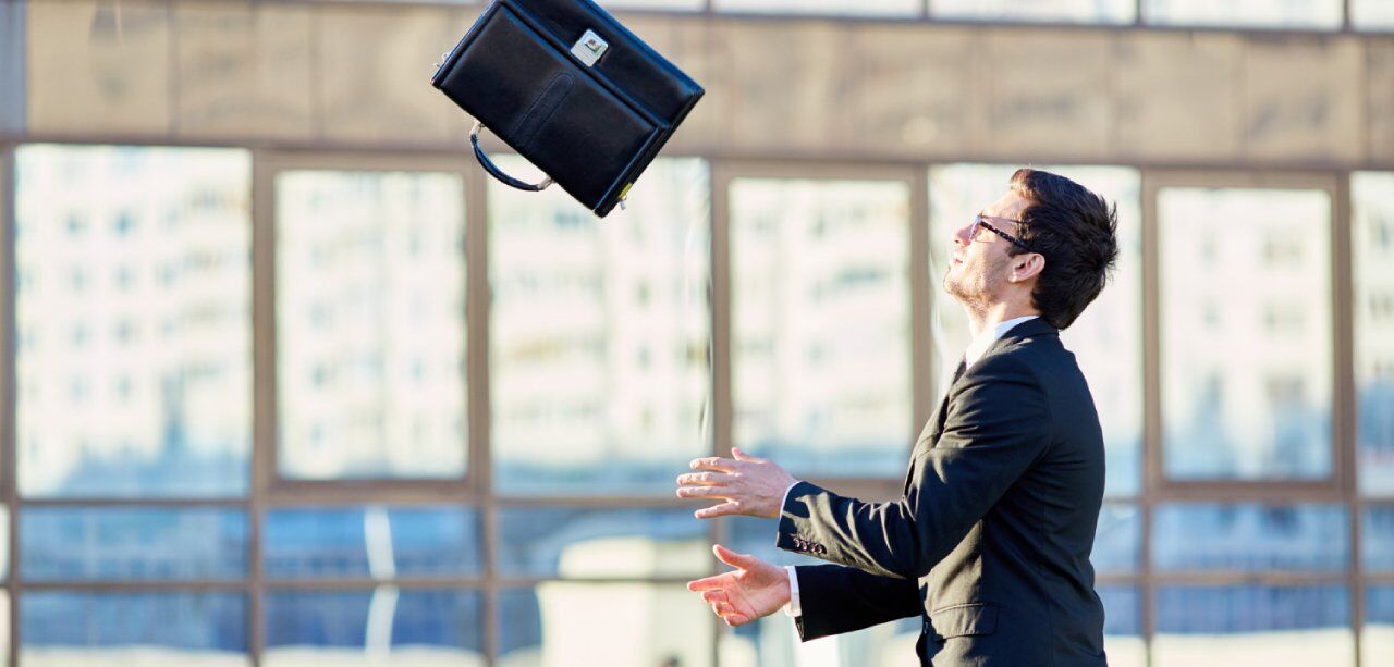 A person in a business suit is outdoors, tossing a black briefcase into the air. The background includes tall, reflective buildings with large windows.