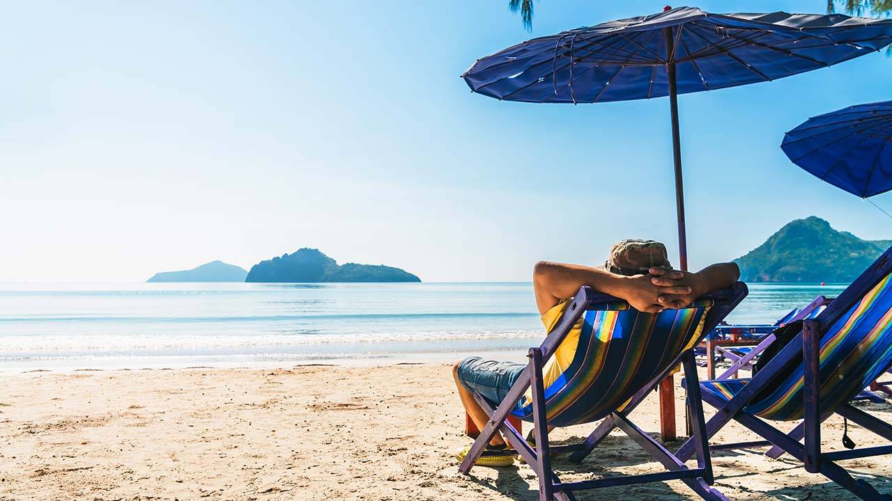 A person relaxes on a colorful beach chair under a large umbrella, facing a serene ocean with distant islands and a clear blue sky. The sandy beach is calm, suggesting a peaceful, sunny day.