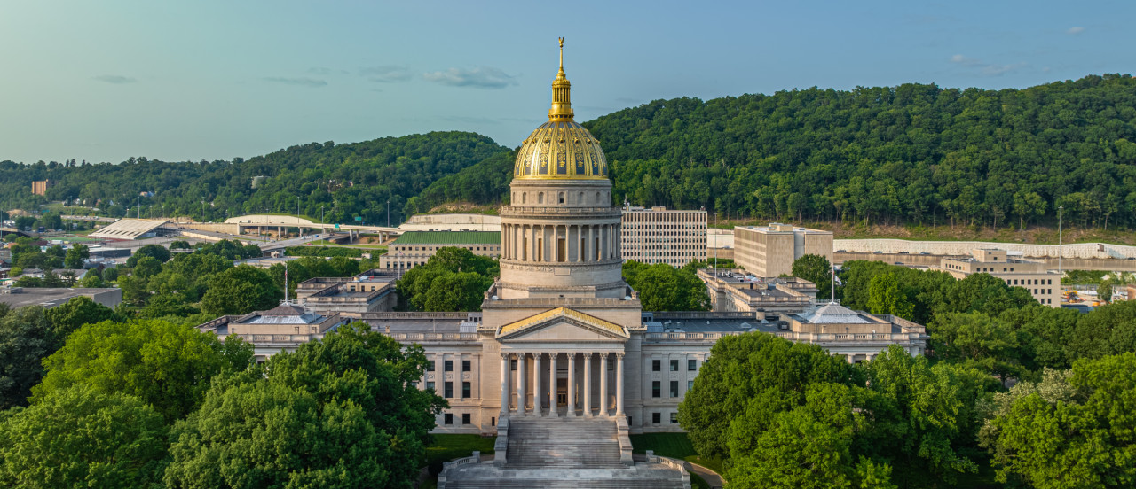 Aerial view of a grand neoclassical building with a prominent golden dome surrounded by trees. The structure is nestled against a backdrop of lush green hills under a clear sky.