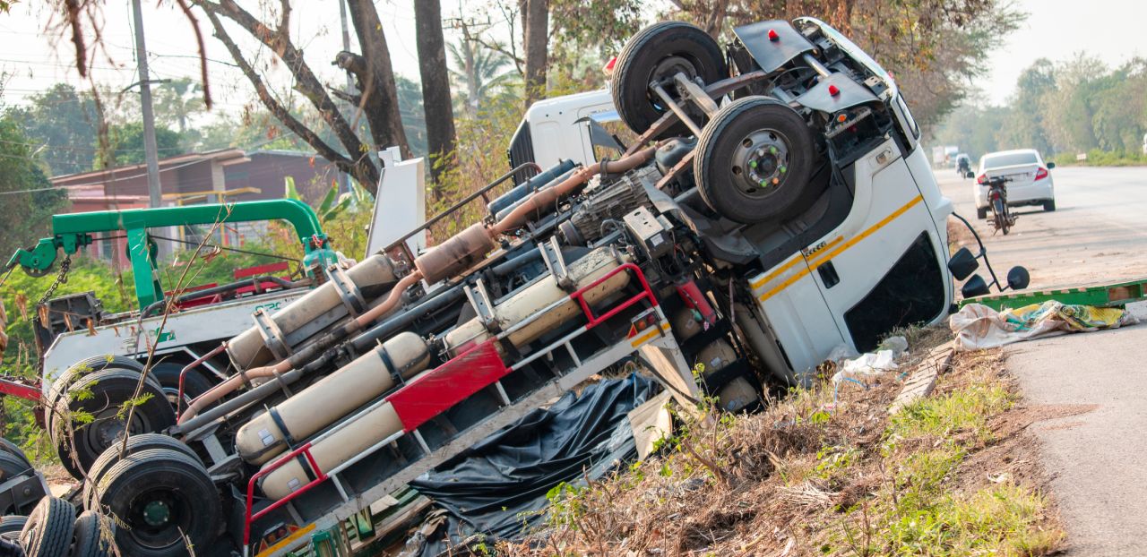 A large white truck is overturned on the side of a rural road, near some trees. The vehicle's undercarriage and wheels are visible, and debris is scattered nearby. Other vehicles and a person on a motorcycle are visible in the background.