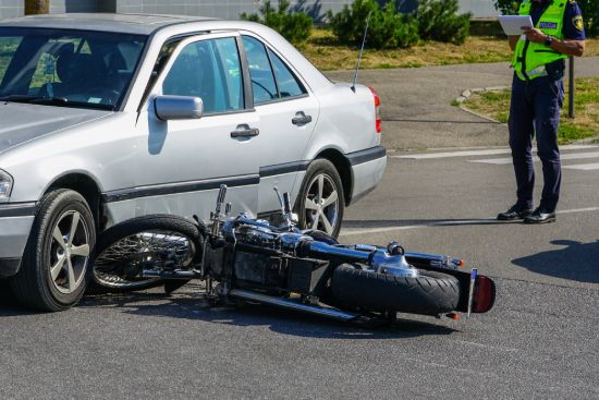 A car and a motorcycle have collided at an intersection. The motorcycle is laying on its side in front of the car. A police officer stands nearby, taking notes.