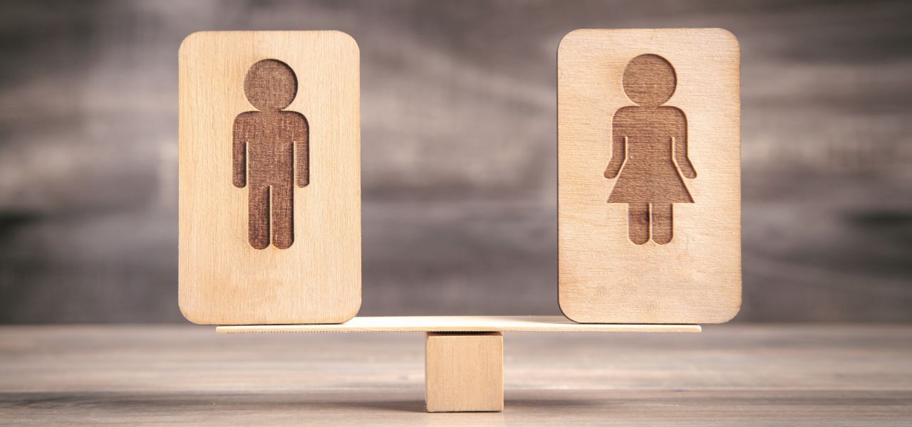 Two wooden blocks on a scale represent gender equality. The left block features a male icon, and the right block features a female icon. The scale is balanced, symbolizing equality.