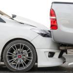 A white sedan has collided with the rear of a gray pickup truck on a road. The sedan's front bumper is crumpled. Both vehicles are stopped, showing visible damage.