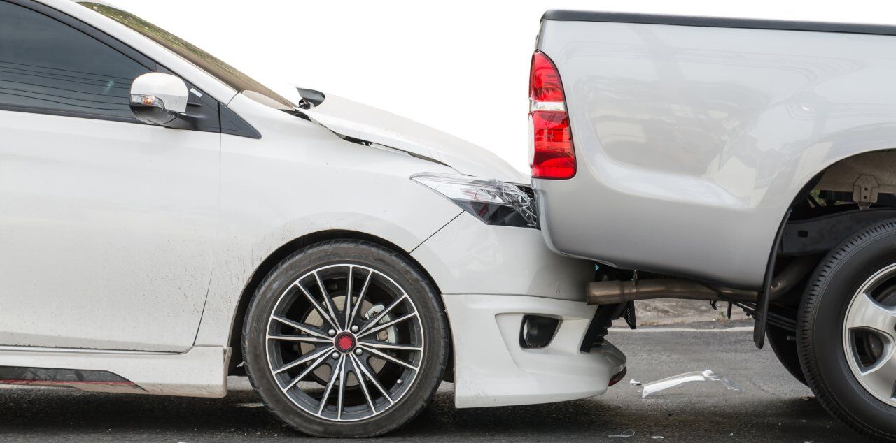 A white sedan has collided with the rear of a gray pickup truck on a road. The sedan's front bumper is crumpled. Both vehicles are stopped, showing visible damage.