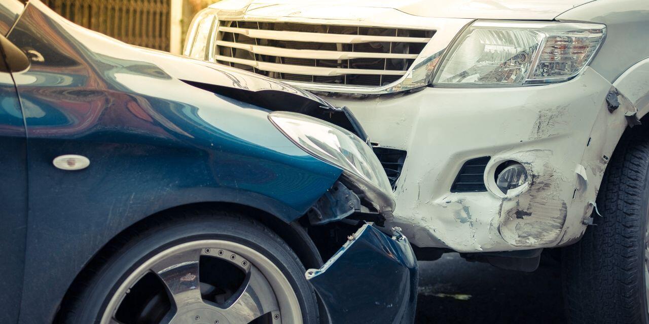 A close-up of two cars with visible damage after a collision. The front of a white car is pressed against the side of a dark blue car, with dents and scratches on their bumpers. The scene appears to be on a street in daylight.