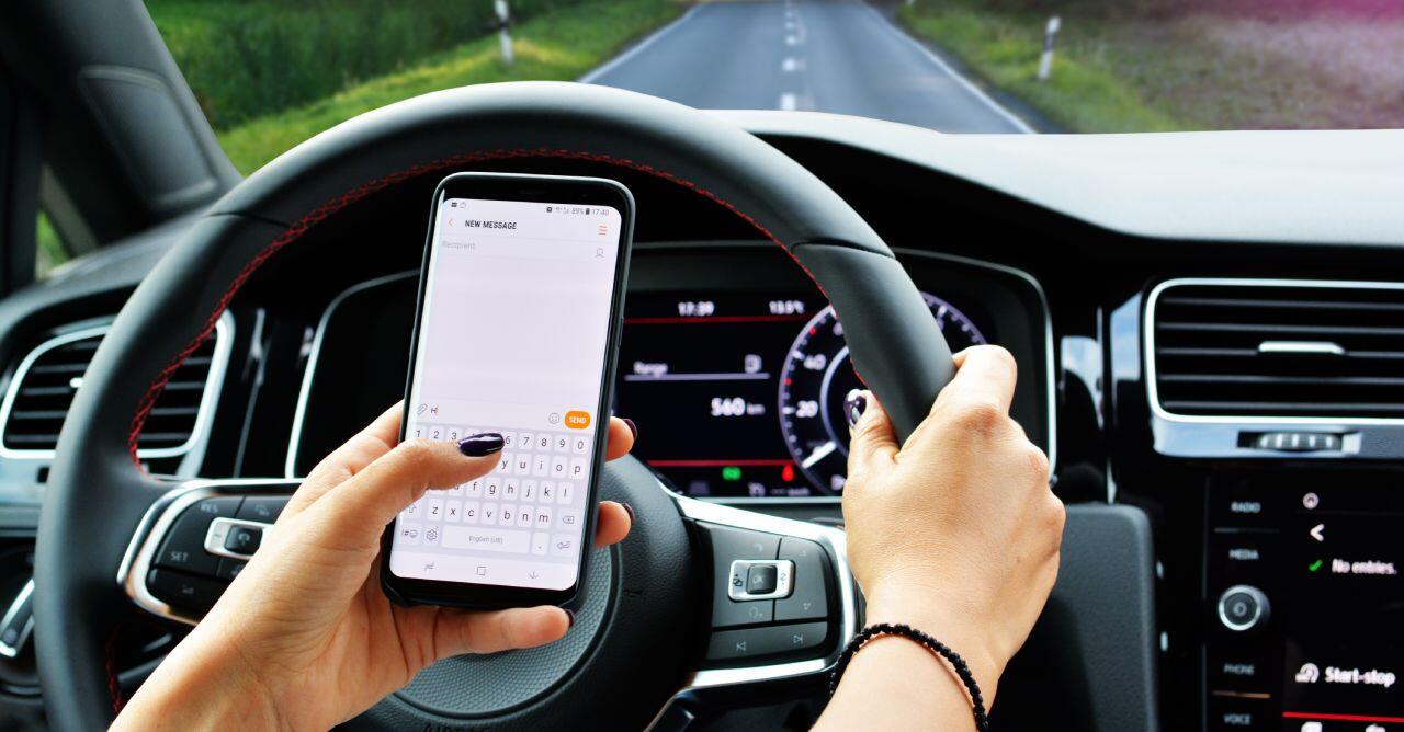 A person is driving a car with one hand on the steering wheel while using a smartphone in the other hand. The road ahead is clear, with trees lining the sides. The car dashboard displays speed and various controls.