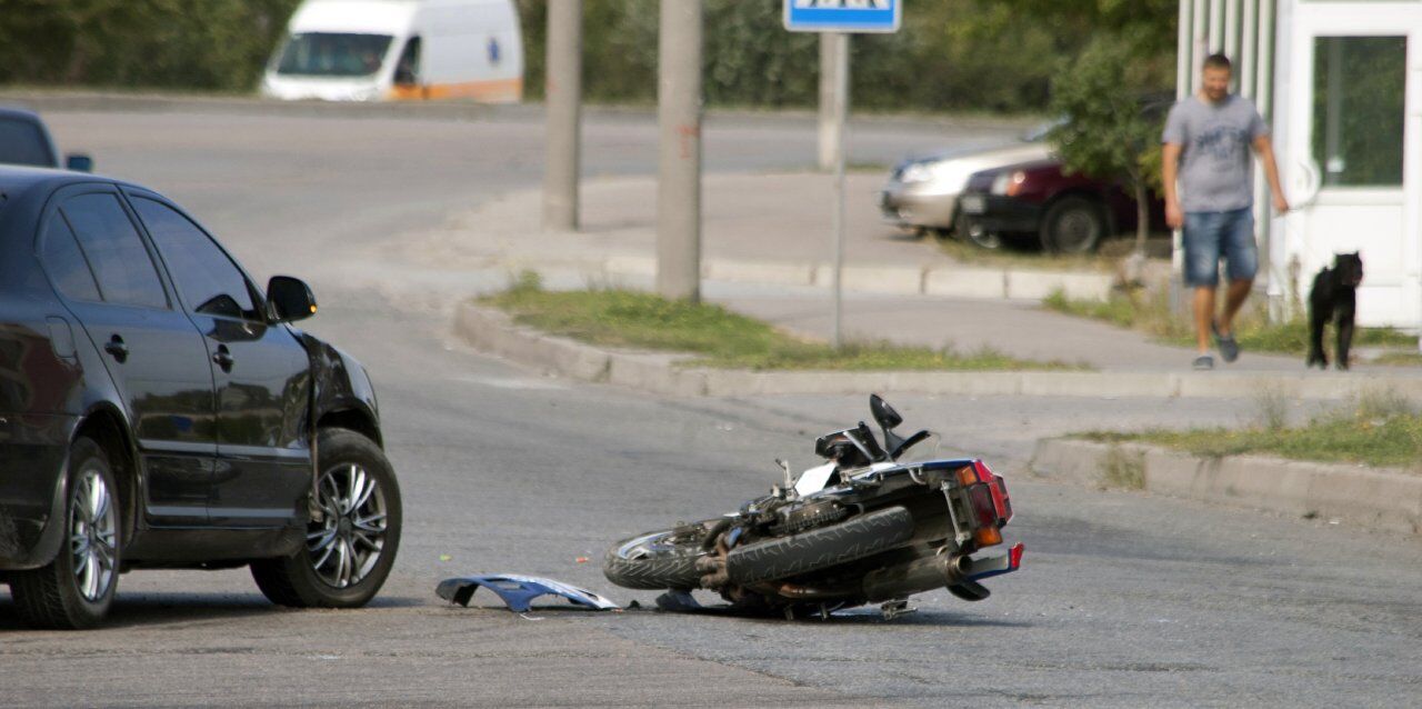 A motorcycle lying on its side in the street near a black car, suggesting an accident. A man and a dog are walking on the sidewalk nearby. An emergency vehicle is visible in the background.