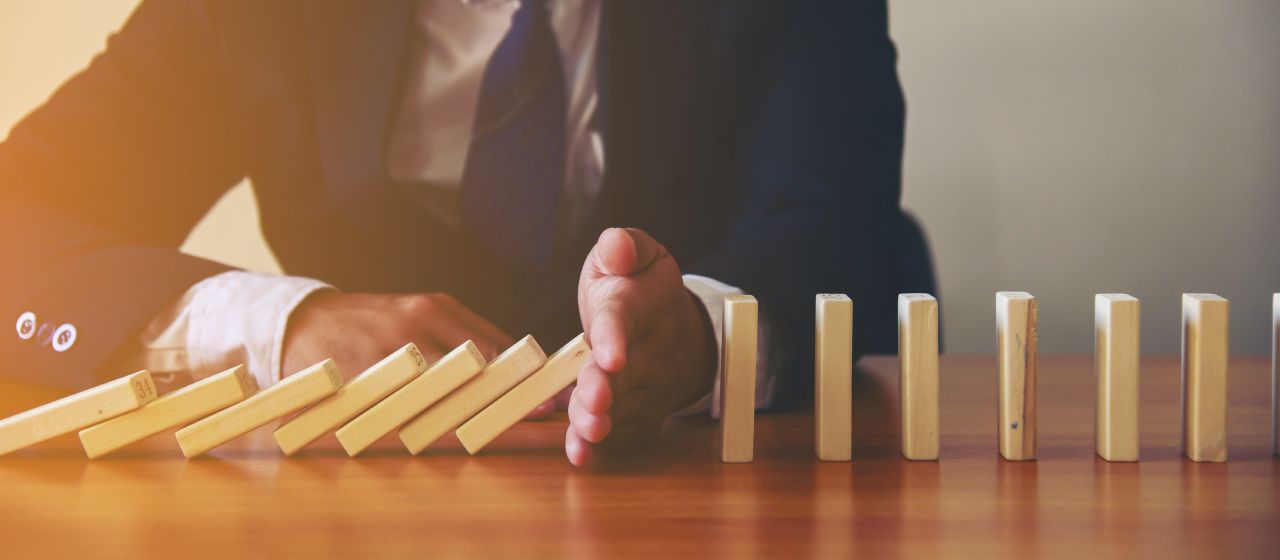 A person in a suit prevents a row of wooden dominoes from falling by placing a hand in their path on a wooden table, suggesting intervention or stopping a chain reaction.