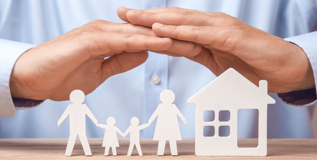 A person's hands are cupped protectively over white wooden cutouts of a family with two adults and two children next to a house figure on a table, symbolizing home and family protection.