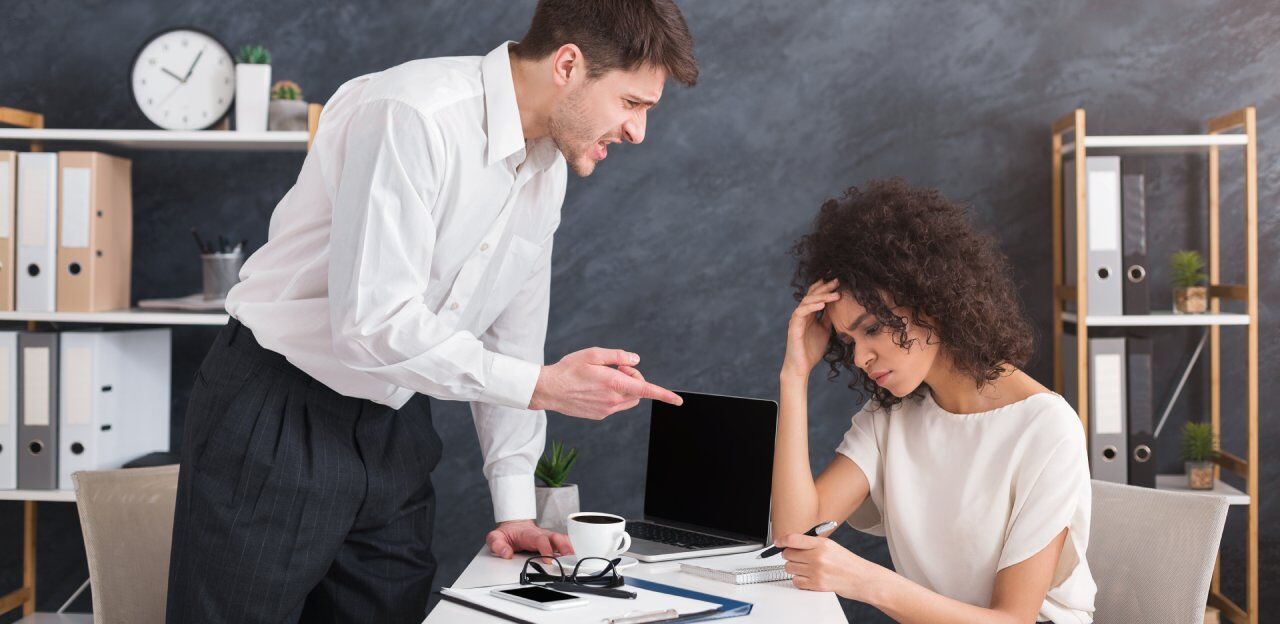 A man stands pointing in frustration at a seated woman who looks stressed, holding her forehead. They are in an office setting with a laptop, a coffee cup, and office supplies on the table. Bookshelves and a clock are in the background.