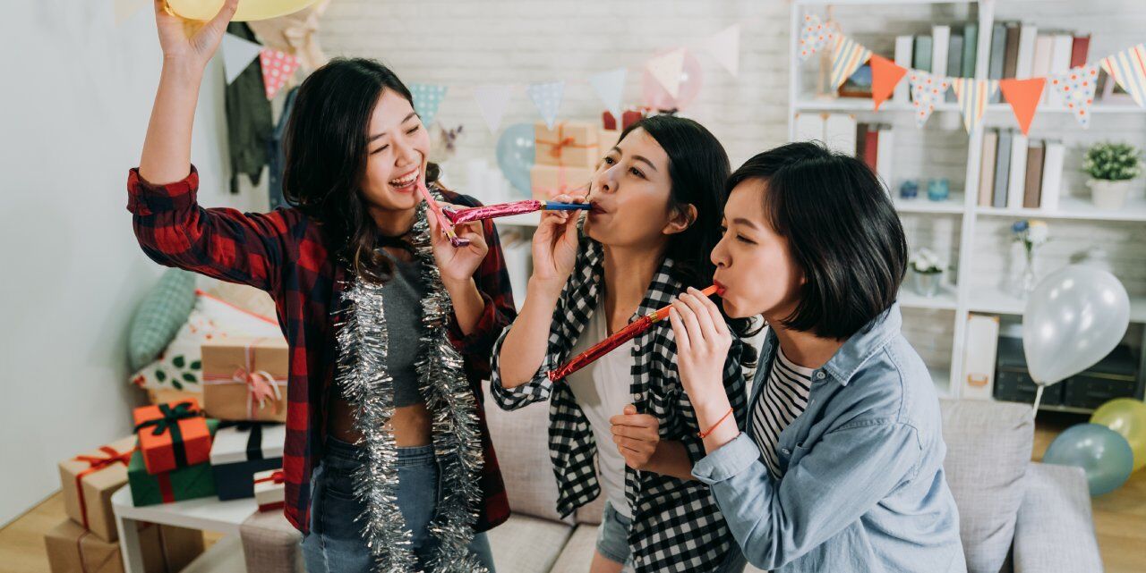 Three women joyfully celebrate indoors, wearing casual clothes. They are blowing party horns, and one has tinsel around her neck. Colorful banners and balloons decorate the background, with wrapped gifts visible on a sofa.