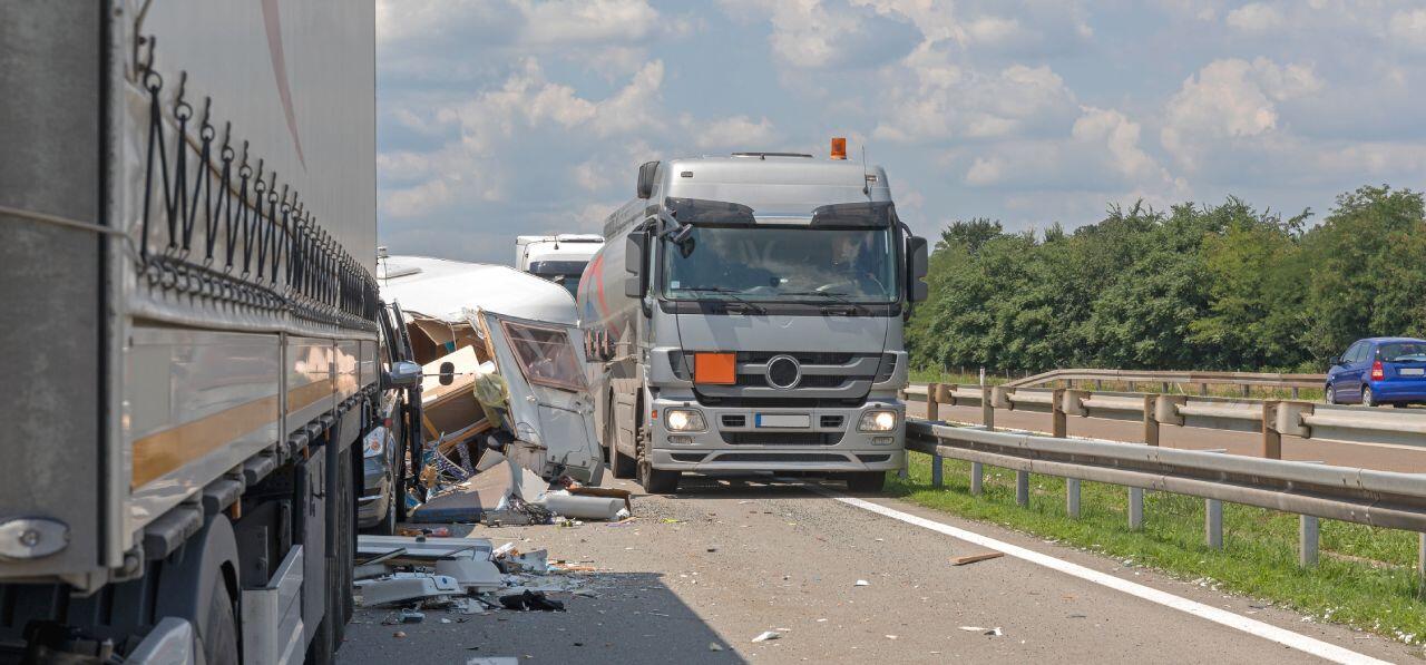A highway accident scene with a damaged truck and a crushed camper on the road. The sky is partly cloudy, and there are vehicles in the background on the other side of the road.