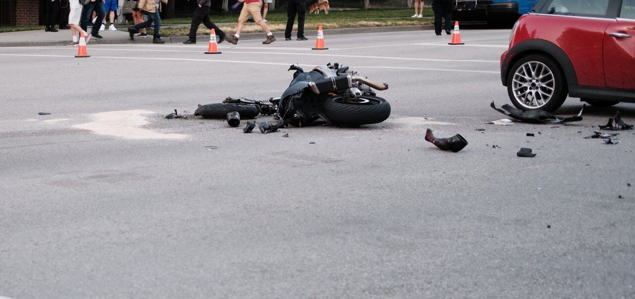 A motorcycle lies damaged on the road near a red car after an accident. Debris is scattered around, and traffic cones block the area. People are walking on the sidewalk in the background.
