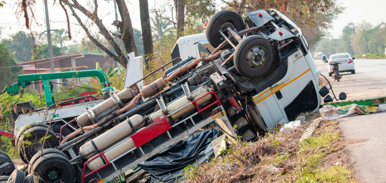 A large truck has flipped onto its side on a roadside, with its wheels in the air. Debris is scattered around, and a motorcyclist rides away in the background. Trees and a small building are visible nearby.