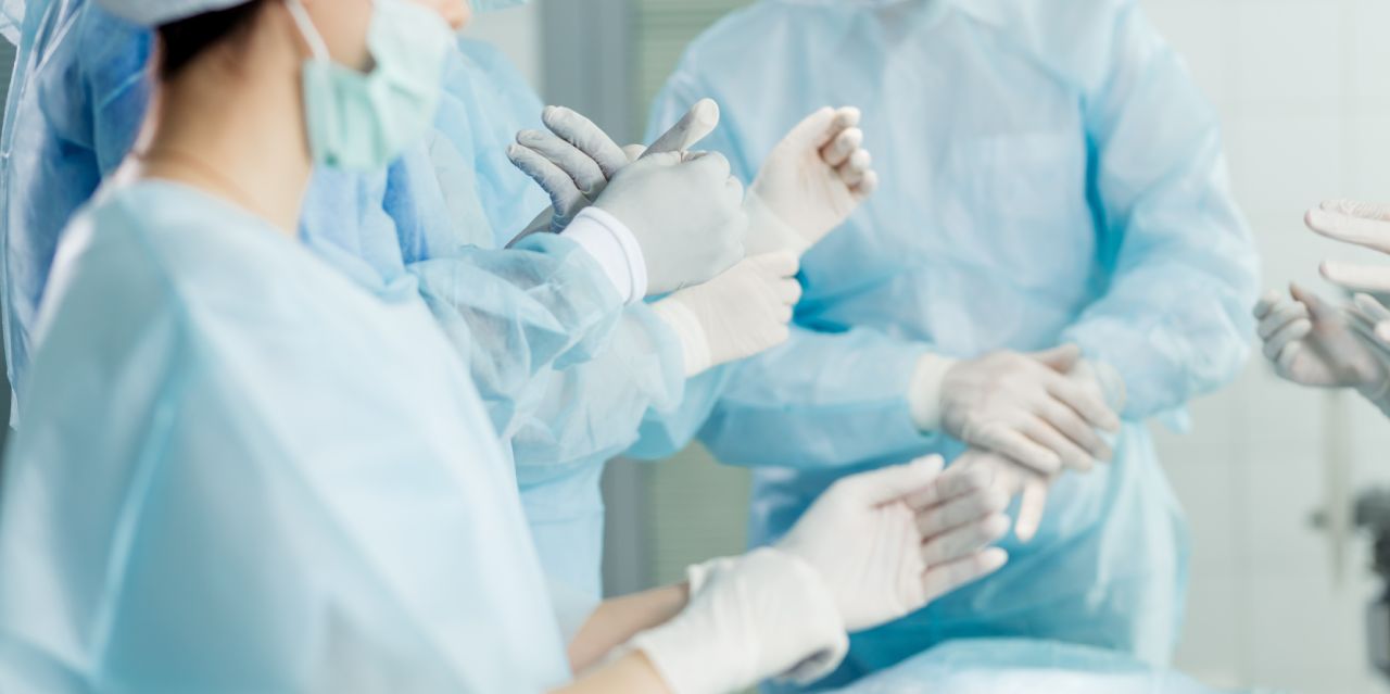 Surgeons in blue surgical gowns and masks gather in an operating room, putting on sterile gloves and preparing for a procedure.