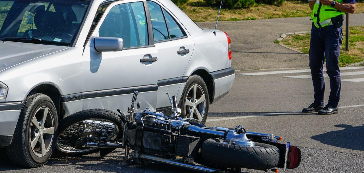 A silver car has collided with a motorcycle, which is lying on its side on the road. A person in a reflective vest stands nearby, possibly assessing the situation or taking notes.