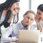 Three women in an office, with two standing and shouting at the third, who is sitting at a desk looking overwhelmed. A laptop and orange coffee mug are on the table. They appear to be in a brightly lit room.