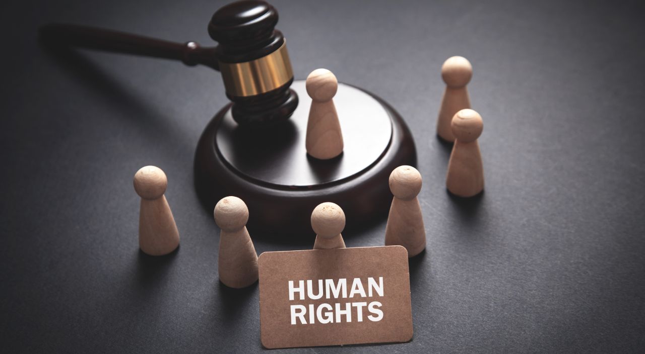 A wooden gavel rests on its base surrounded by wooden pawn figures. In the foreground, a brown card with white letters reads "HUMAN RIGHTS," suggesting the theme of justice and human rights advocacy.