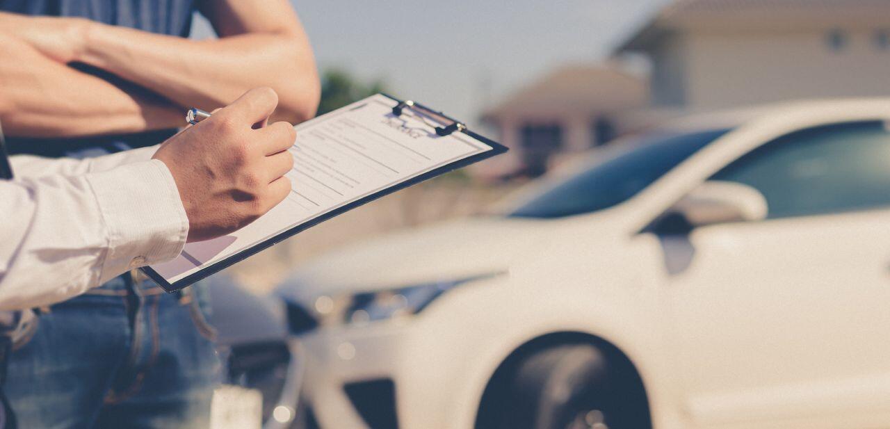 A person holding a clipboard is writing on a document, while another person stands beside them with crossed arms. In the background, a white car is parked on a sunny day.