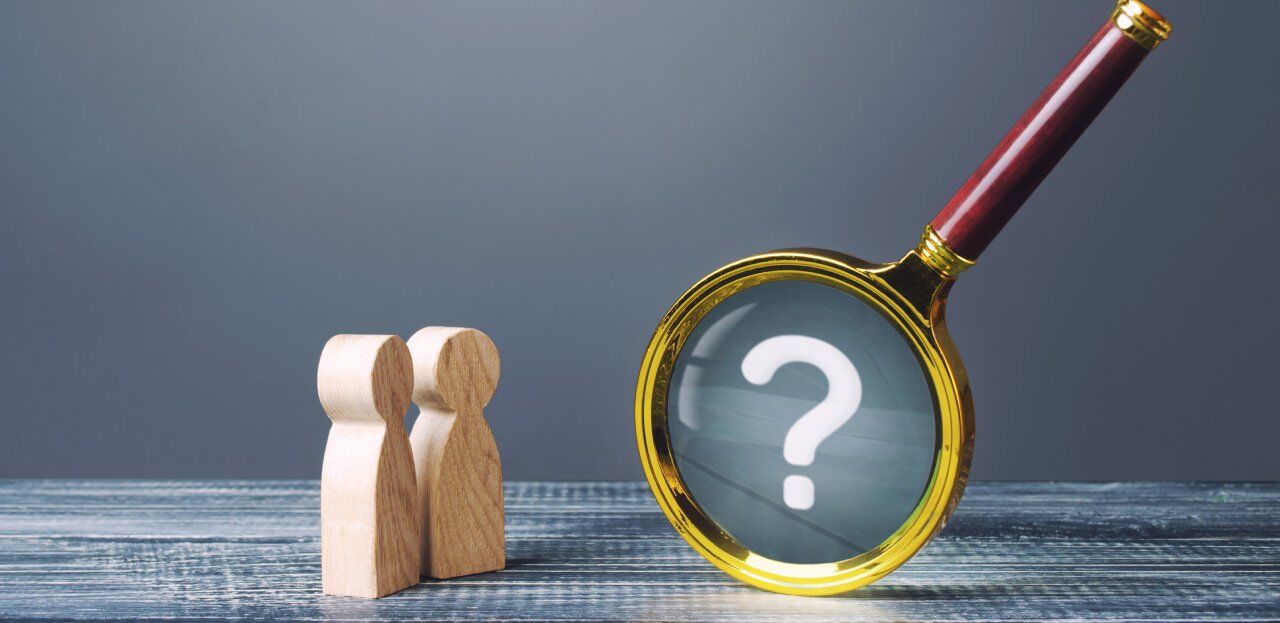 Two wooden figurines stand on a wooden surface, next to a large magnifying glass with a golden rim. The magnifying glass highlights a white question mark on a gray background.