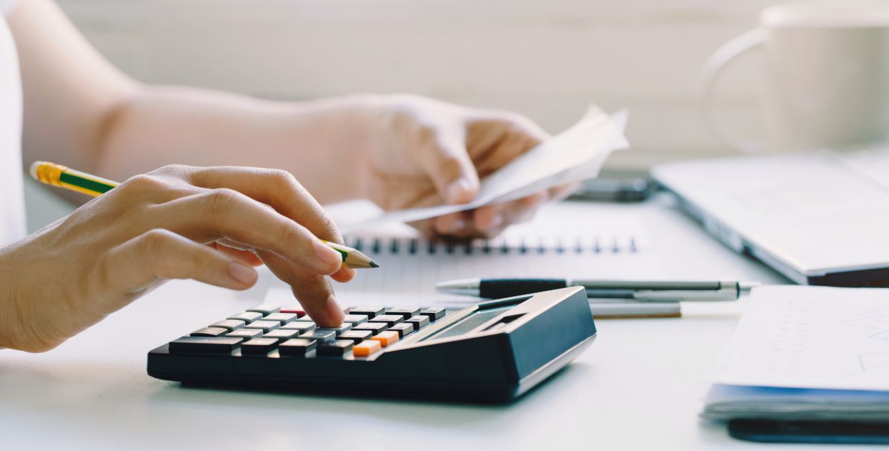 Close-up of a person's hands using a calculator and holding a piece of paper. A notebook, pen, and open laptop are on the desk. A coffee mug is slightly visible in the background, suggesting a workspace or office setting.