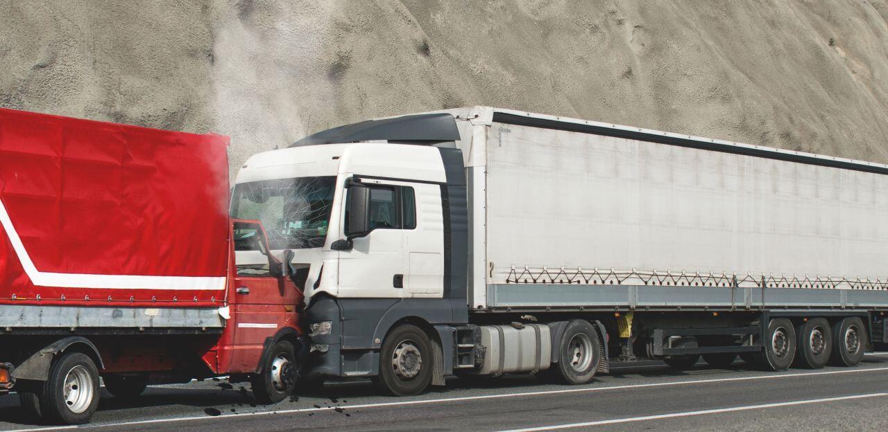 A white semi-truck is crashed into the rear of a red truck on a highway, with a dirt embankment visible in the background. Smoke appears to be rising from the front of the white truck.