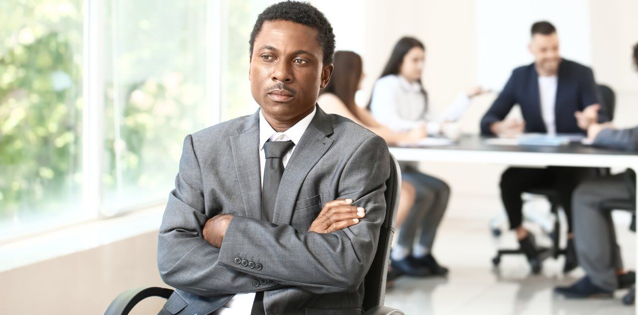 A man in a gray suit sits with arms crossed and looks contemplative. He is in an office setting, with four colleagues blurred in the background, engaged in conversation at a table. Large windows let in natural light.