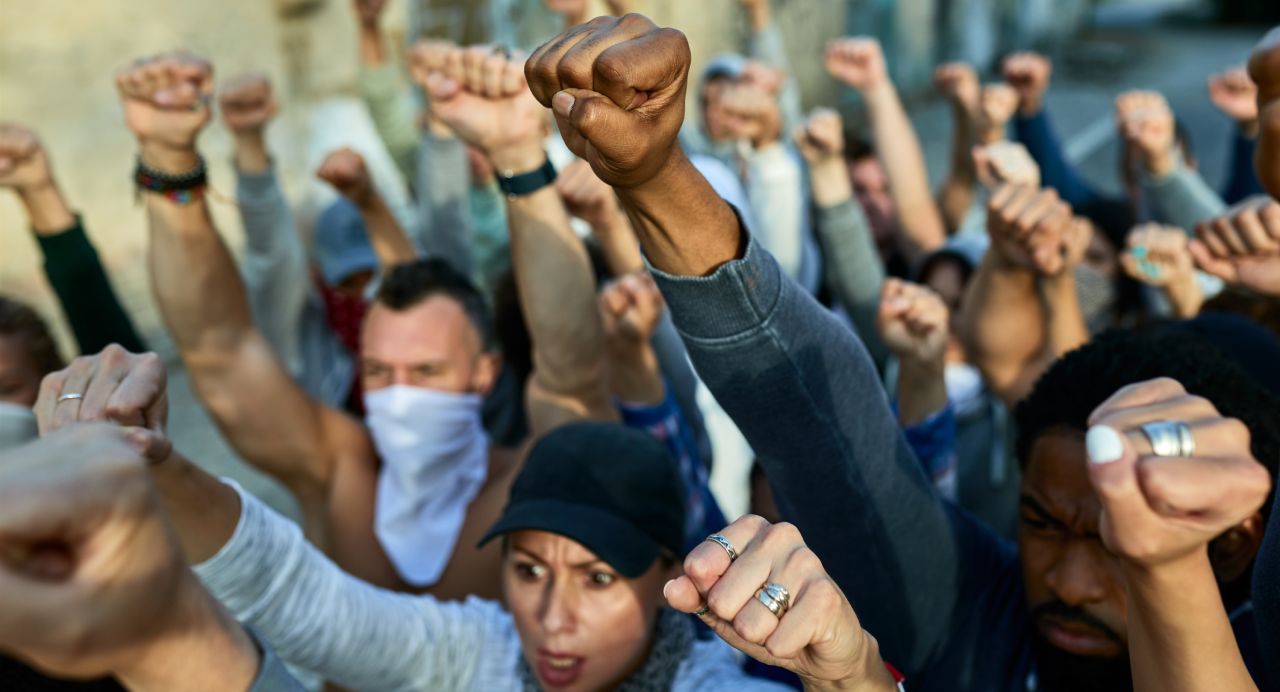 A diverse group of people stand closely together with raised fists, expressing solidarity and protest. The setting appears to be outdoors, and their expressions are serious and determined. Some individuals wear face coverings.