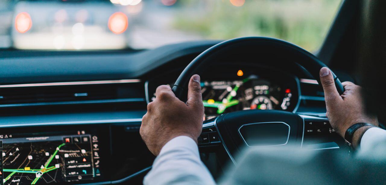 Close-up of a person driving a car, holding the steering wheel with both hands. The dashboard displays illuminated dials and a digital navigation screen. The road ahead is blurry, suggesting motion and focus on driving.