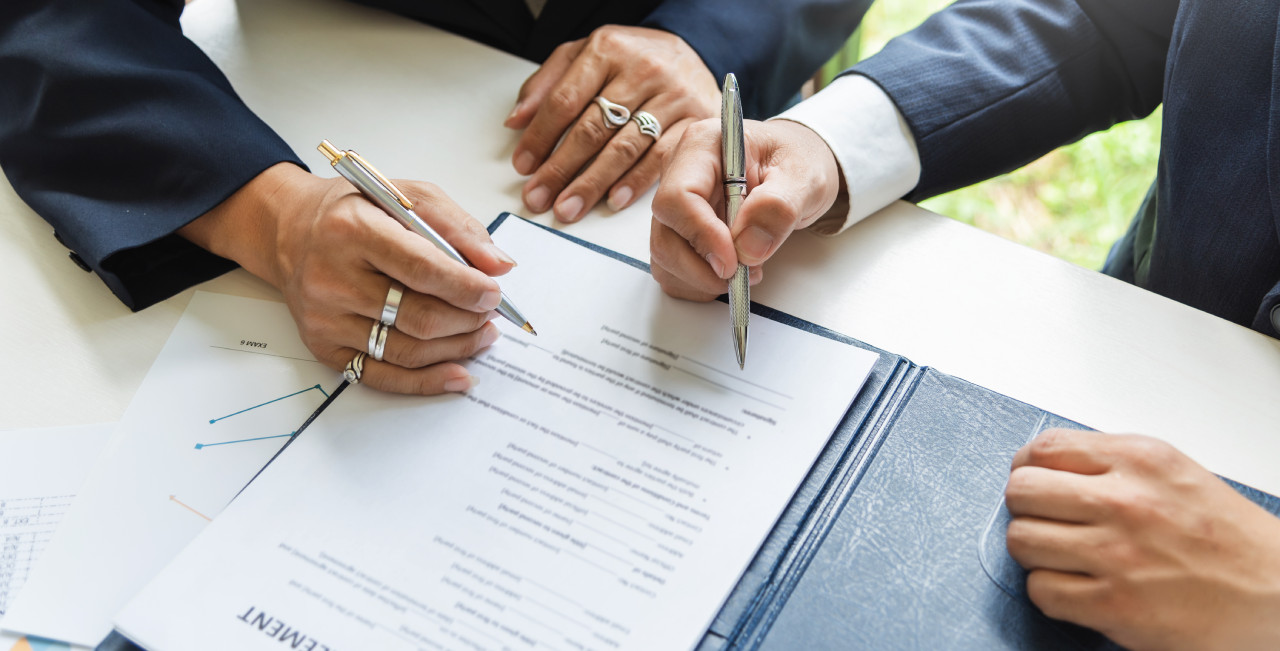 Two people in business attire are signing a document placed on a table. One person holds a silver pen, and the other a black pen. Nearby, a blue folder and some charts on white paper are visible. The setting appears professional.