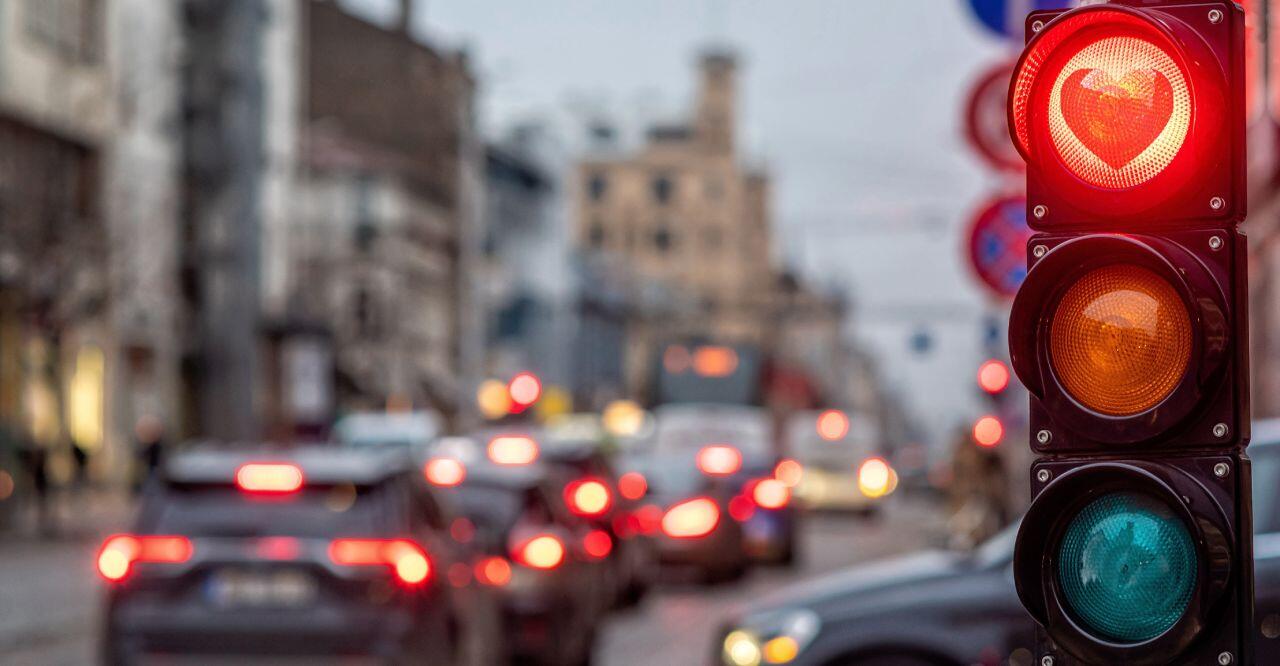 A city street scene at dusk with cars in traffic and a traffic light displaying red. The background is blurred, with buildings and street signs visible.