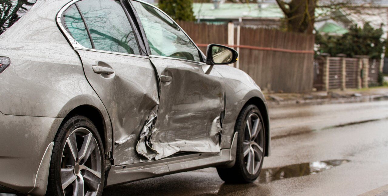 A silver car with visible damage on the driver's side door is parked on a wet street. The surrounding area features a wooden fence and some trees, indicating a residential neighborhood.